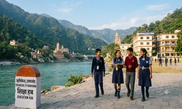 Four students in school uniforms walking along a paved path by the Ganges River in Rishikesh, Uttarakhand, with temples, mountains, and school buildings in the background.