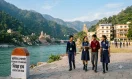 Four students in school uniforms walking along a paved path by the Ganges River in Rishikesh, Uttarakhand, with temples, mountains, and school buildings in the background.